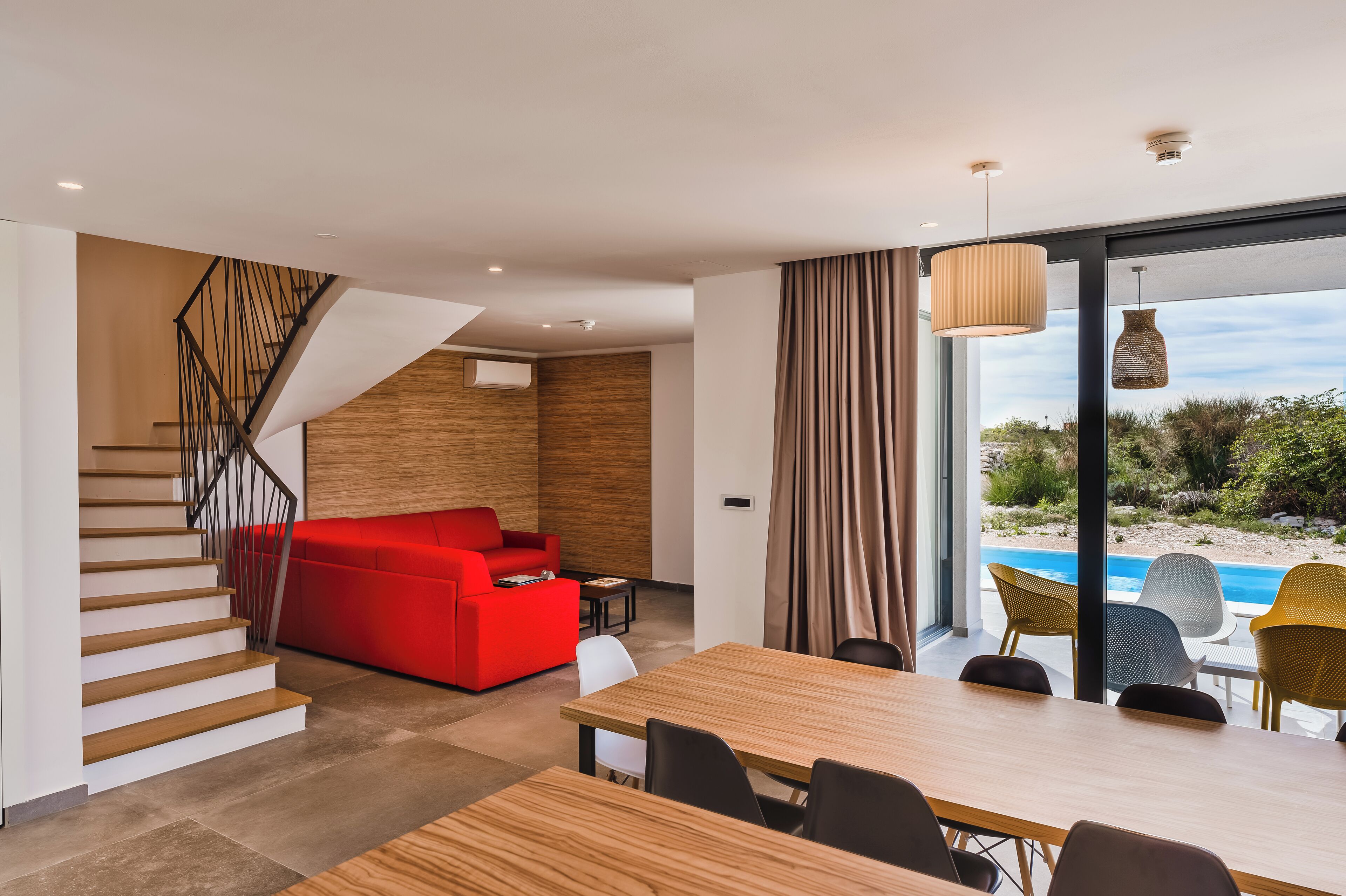 A modern interior with a staircase, red sofa, dining table, and a view of a pool.