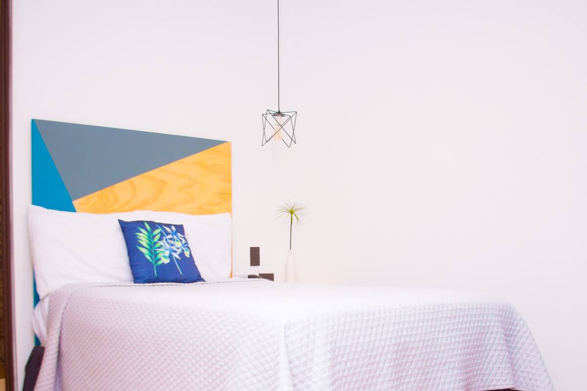 Een netjes opgemaakt bed met een witte spreidek en een paarse onderrok, een geometrisch kunstwerk erboven, en een hanglamp.
