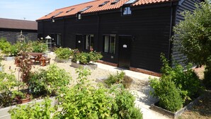 A black building with a tiled roof, multiple windows, and a garden area with plants and a wooden table. - Mollett's Farm (Saxmundham)
