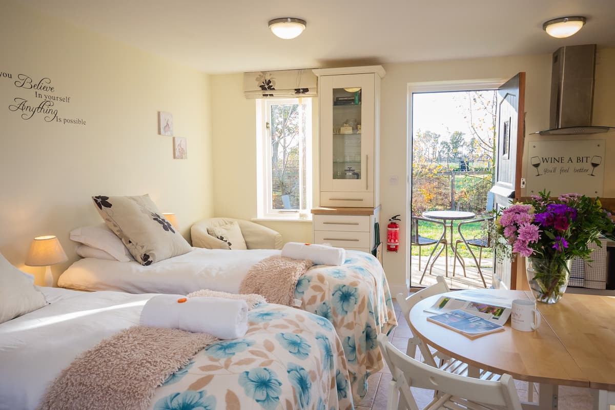 Een gezellige slaapkamer met twee bedden, een eettafel met een bloemenvaas, en een keukenruimte zichtbaar door een open deur.