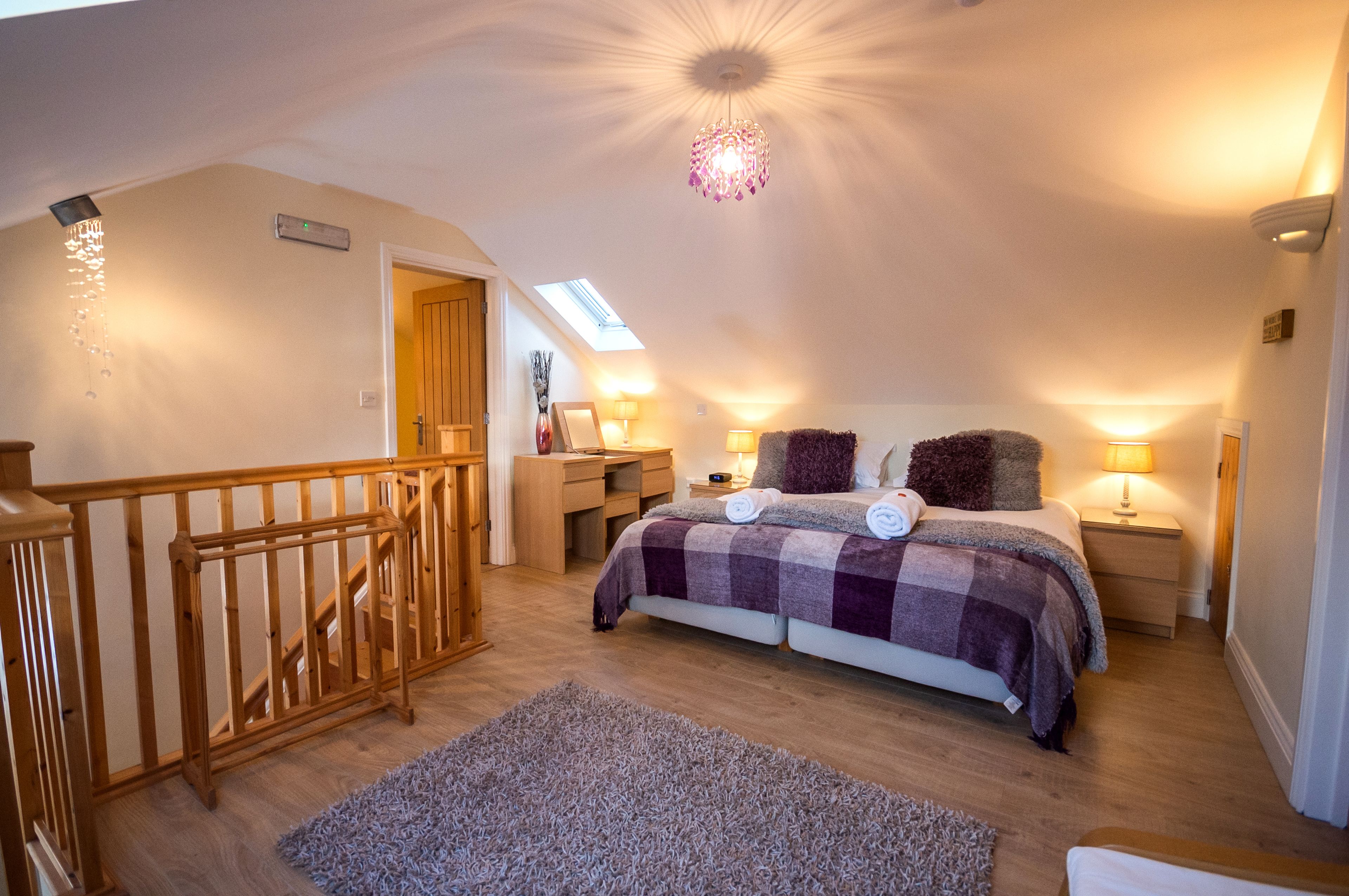 A bedroom with a bed, a desk, a chair, a wooden railing, a skylight, and a chandelier.
