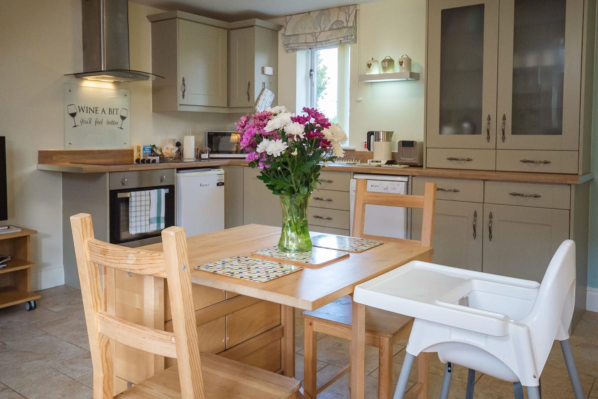 Een keuken met houten kastjes, een eettafel met stoelen, een witte kinderstoel en een vaas met bloemen.