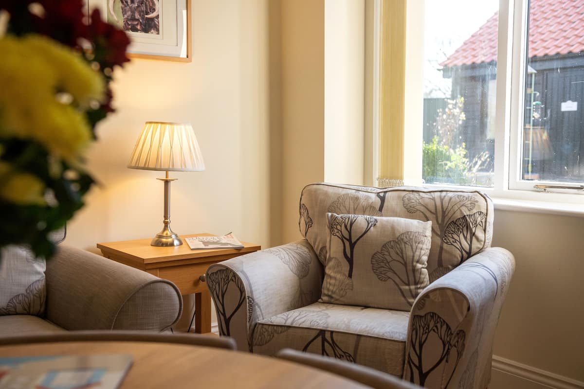 Een gezellige woonkamer met een bank en fauteuils, een bijzettafeltje met een lamp, en een raam met uitzicht op een huis.