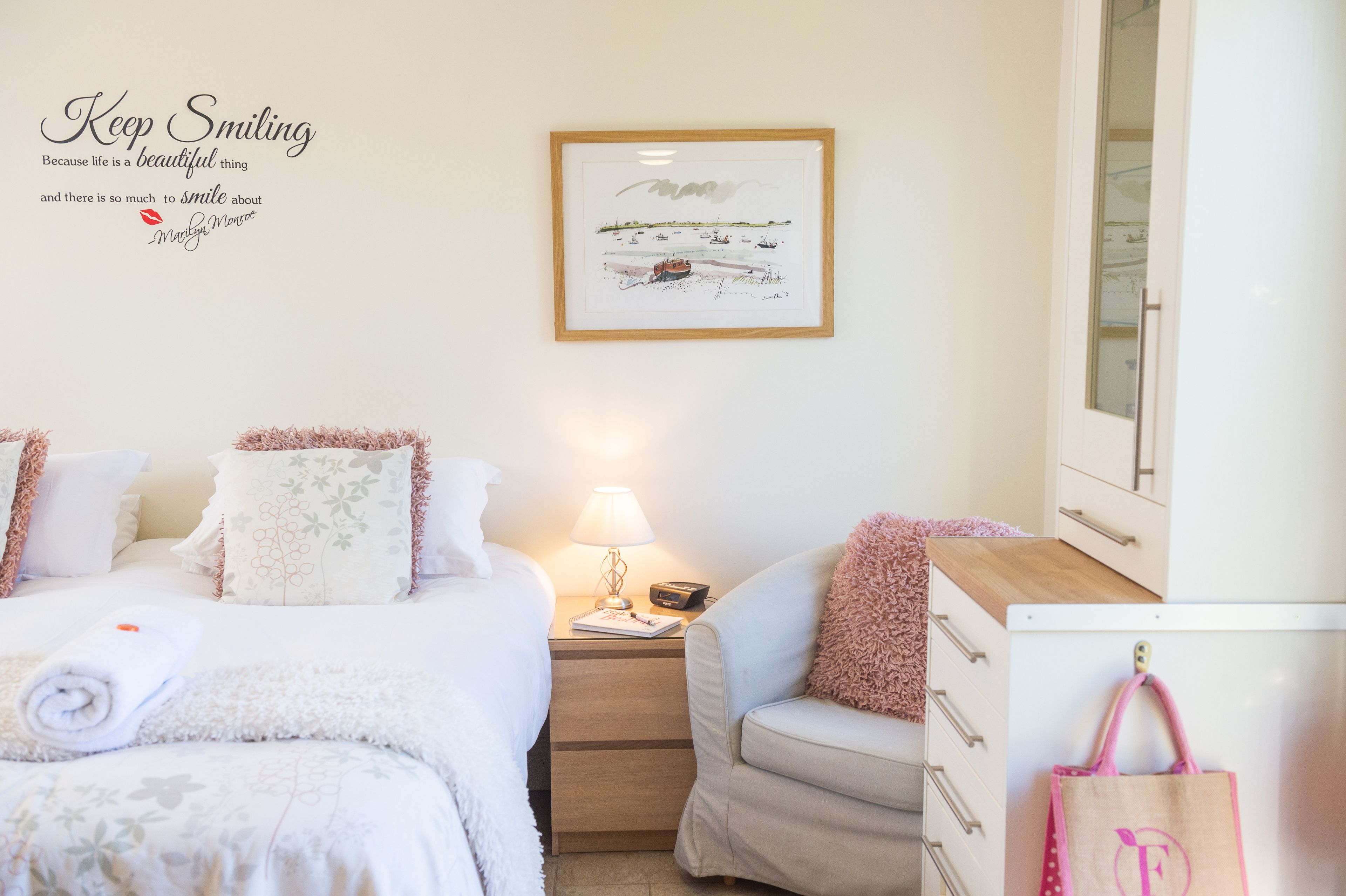 A neatly arranged hotel room with a bed, a chair, a nightstand with a lamp, and a framed quote on the wall.
