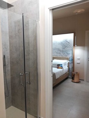 Un baño moderno con ducha envuelta en vidrio y una cama a la vista con colchón y almohadas blancas en la habitación contigua.