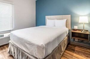 Una cama bien hecha con una colcha blanca y almohadas, un cabecero de madera, una mesita de noche con una lámpara y una ventana con persianas.