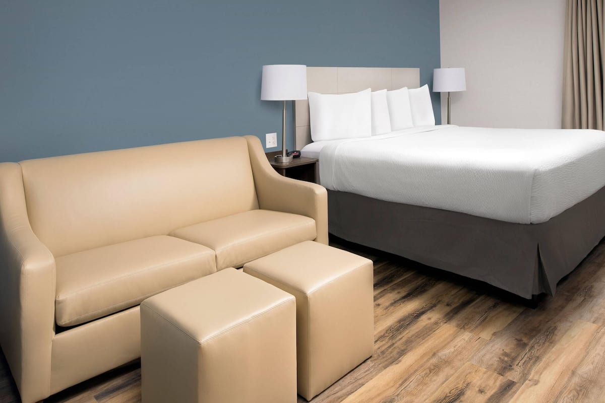 A hotel room with a beige sofa, a matching ottoman, a bed with white linens, and a nightstand with two lamps.