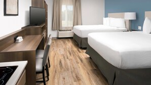 Une chambre d’hôtel avec un bureau en bois, un téléviseur à écran plat, un lit avec du linge de lit blanc, une table de chevet avec une lampe et une fenêtre avec des rideaux.