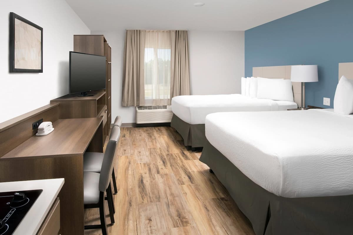 A hotel room with a wooden desk, a flat-screen TV, a bed with white linens, a bedside table with a lamp, and a window with curtains.