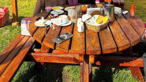 A family dining at a wooden picnic table under a metal roof, with a grassy field and hills in the background. - Tambo Lodge Canta (Canta)