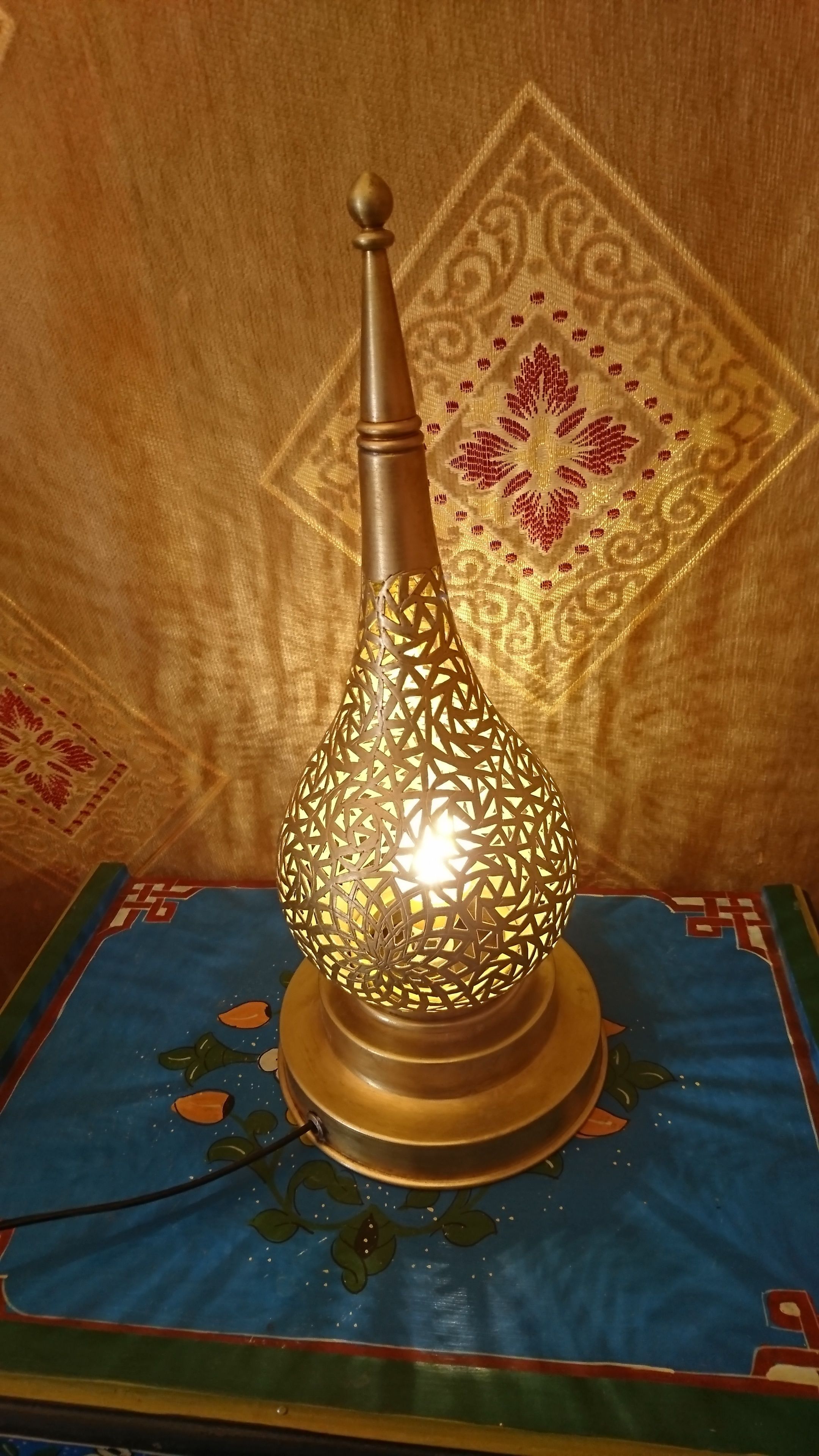 A decorative lamp with a geometric pattern on a table with a blue cloth and a patterned table runner.