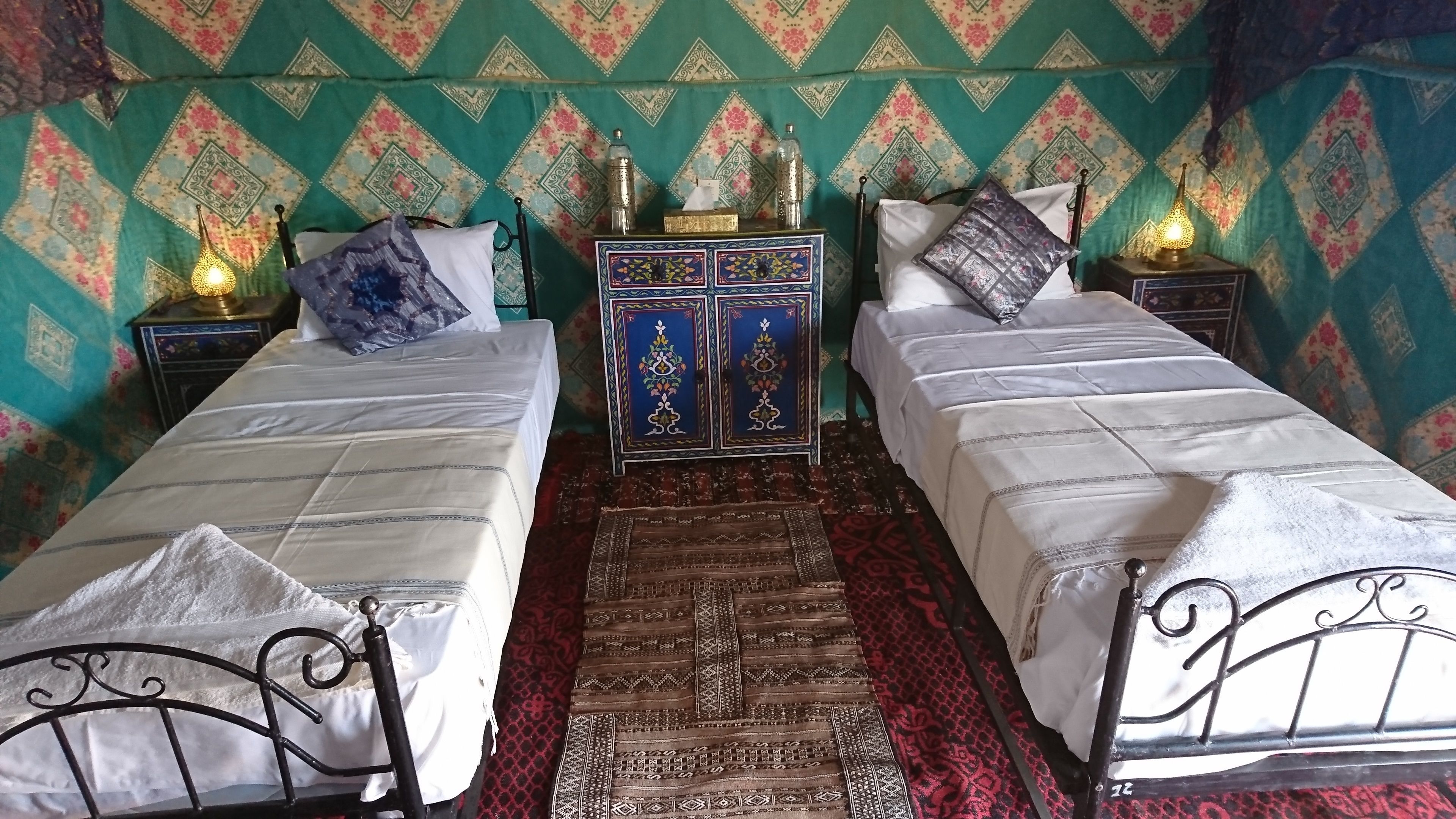 Une chambre avec deux lits simples, un tapis à motifs, une commode décorative et une table de chevet avec une lampe.