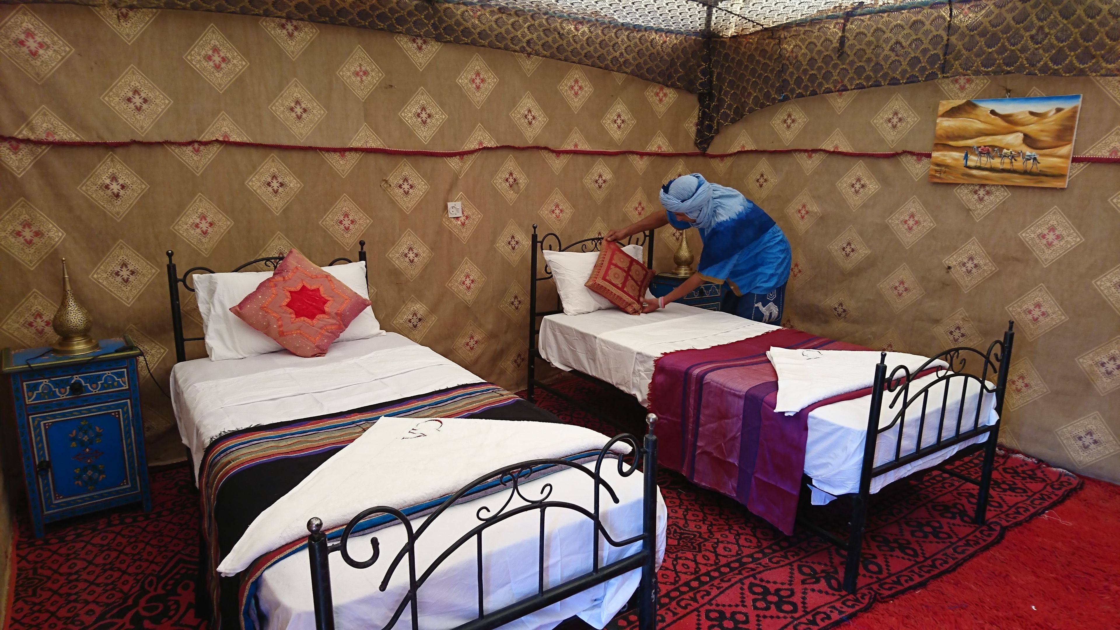 A room with two single beds, a nightstand with a lamp, patterned wallpaper, and a poster on the wall.