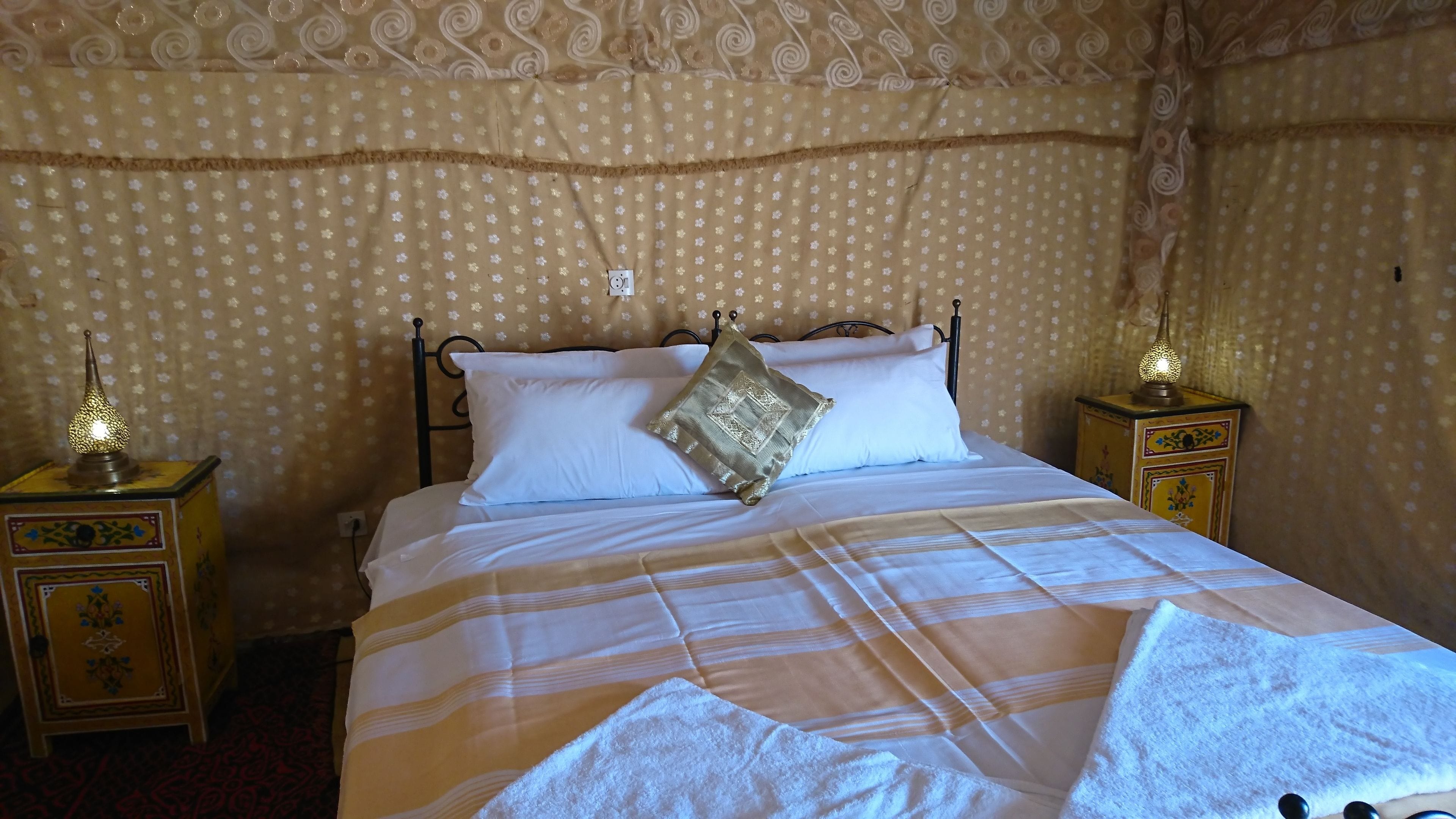 Cama com roupa branca, manta listrada e almofada decorativa. Há duas mesas de cabeceira com luminárias.