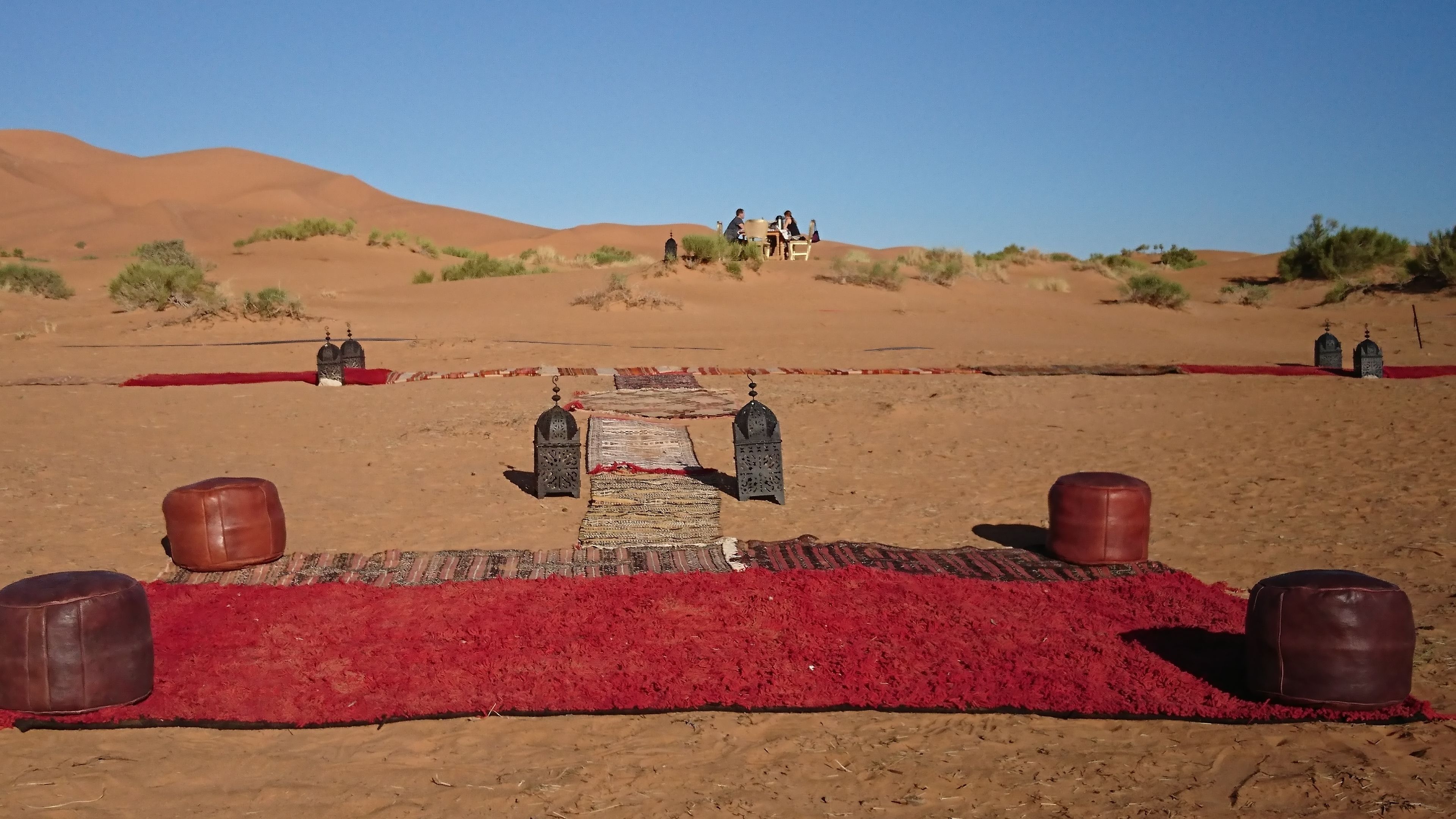 Paisaje desértico con una alfombra roja que conduce a una estructura central, posiblemente un punto de encuentro o entrada, rodeada de dunas de arena y vegetación escasa.