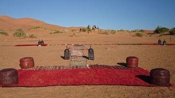 Paisaje desértico con una alfombra roja que conduce a una estructura central, posiblemente un punto de encuentro o entrada, rodeada de dunas de arena y vegetación escasa.