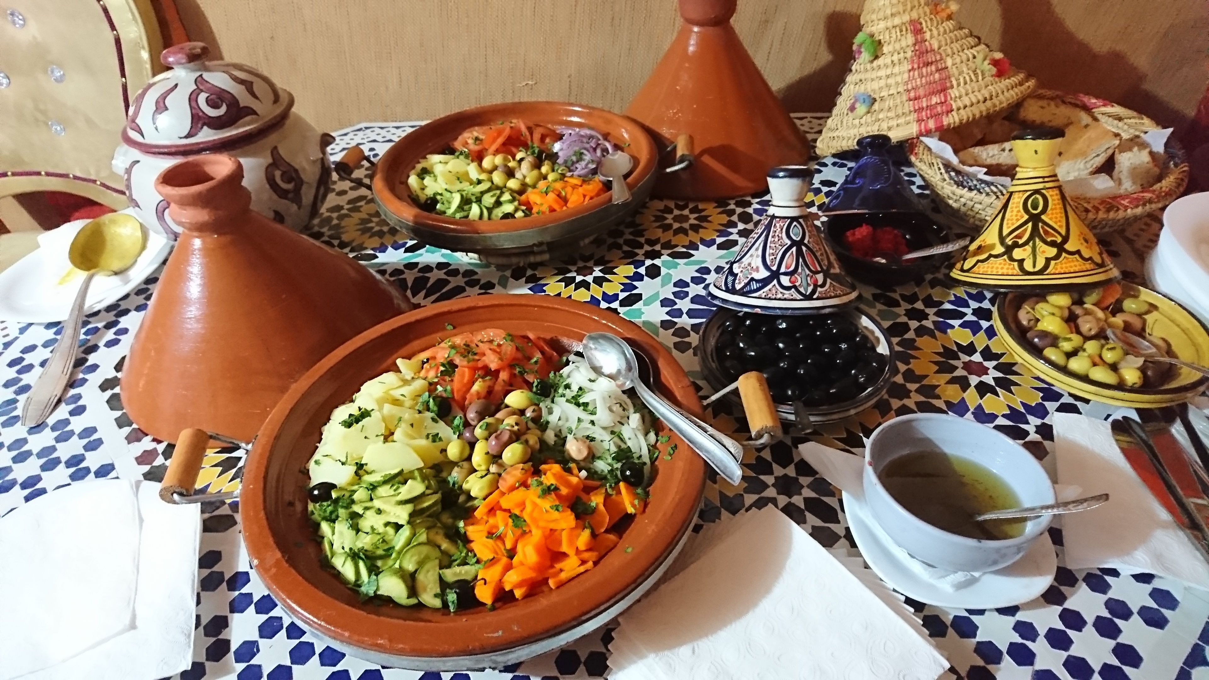 A traditional Moroccan dining setup with a variety of dishes, including couscous, vegetables, and olives, served on terracotta plates and surrounded by ornate Moroccan tea pots and a patterned tablecloth.