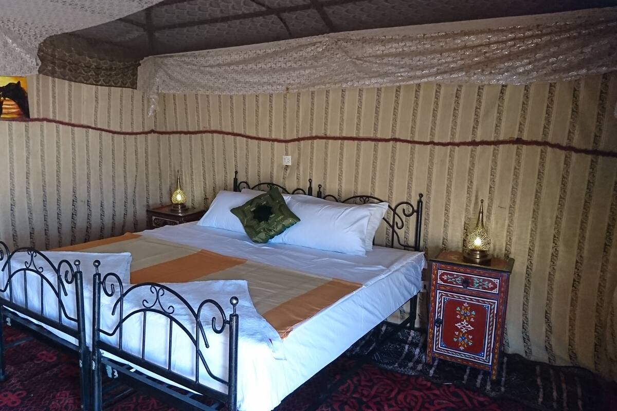 Un lit avec un cadre en métal, une literie blanche et un oreiller vert. Il y a deux tables de chevet avec des lampes. La pièce est décorée de papier peint à rayures et recouverte d’un tapis à motifs.