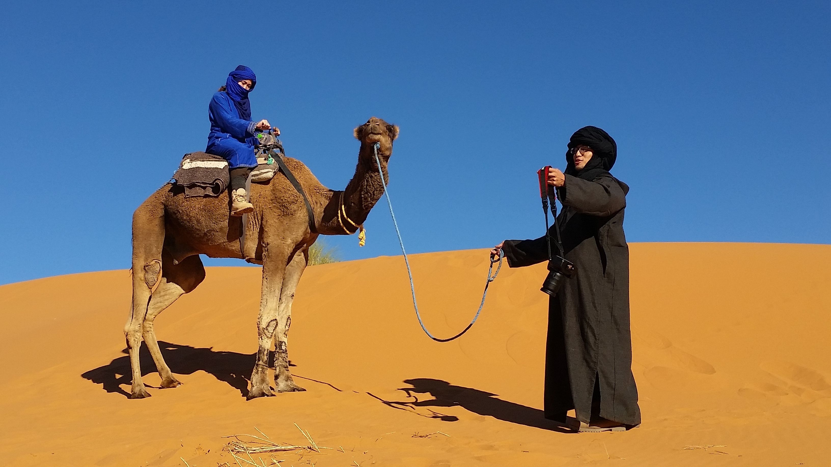 A camel with a rider in blue attire and a person in black attire holding a rope.