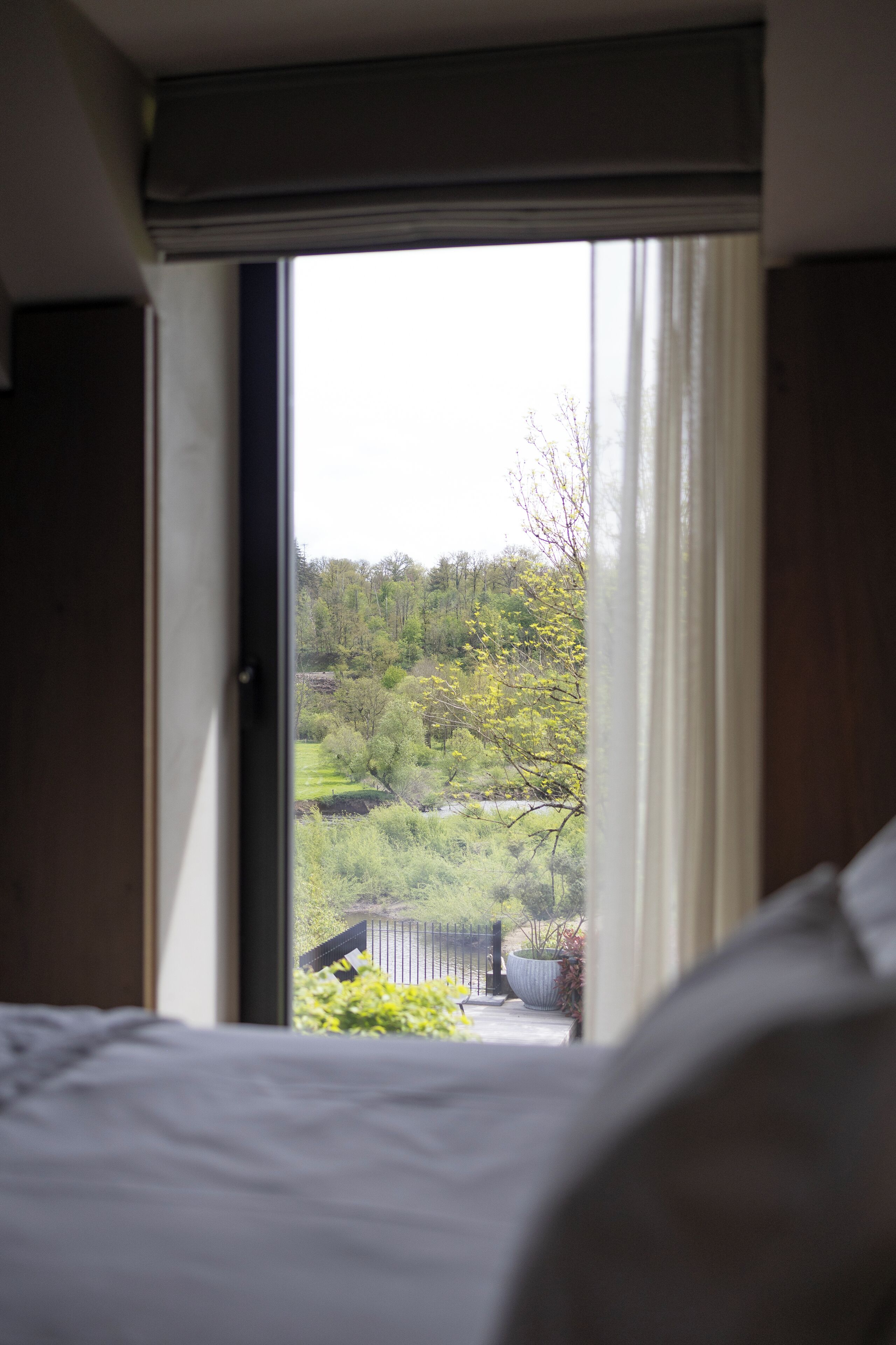 A bedroom with a large window, a bed, and a view of a green landscape with trees and a fence.