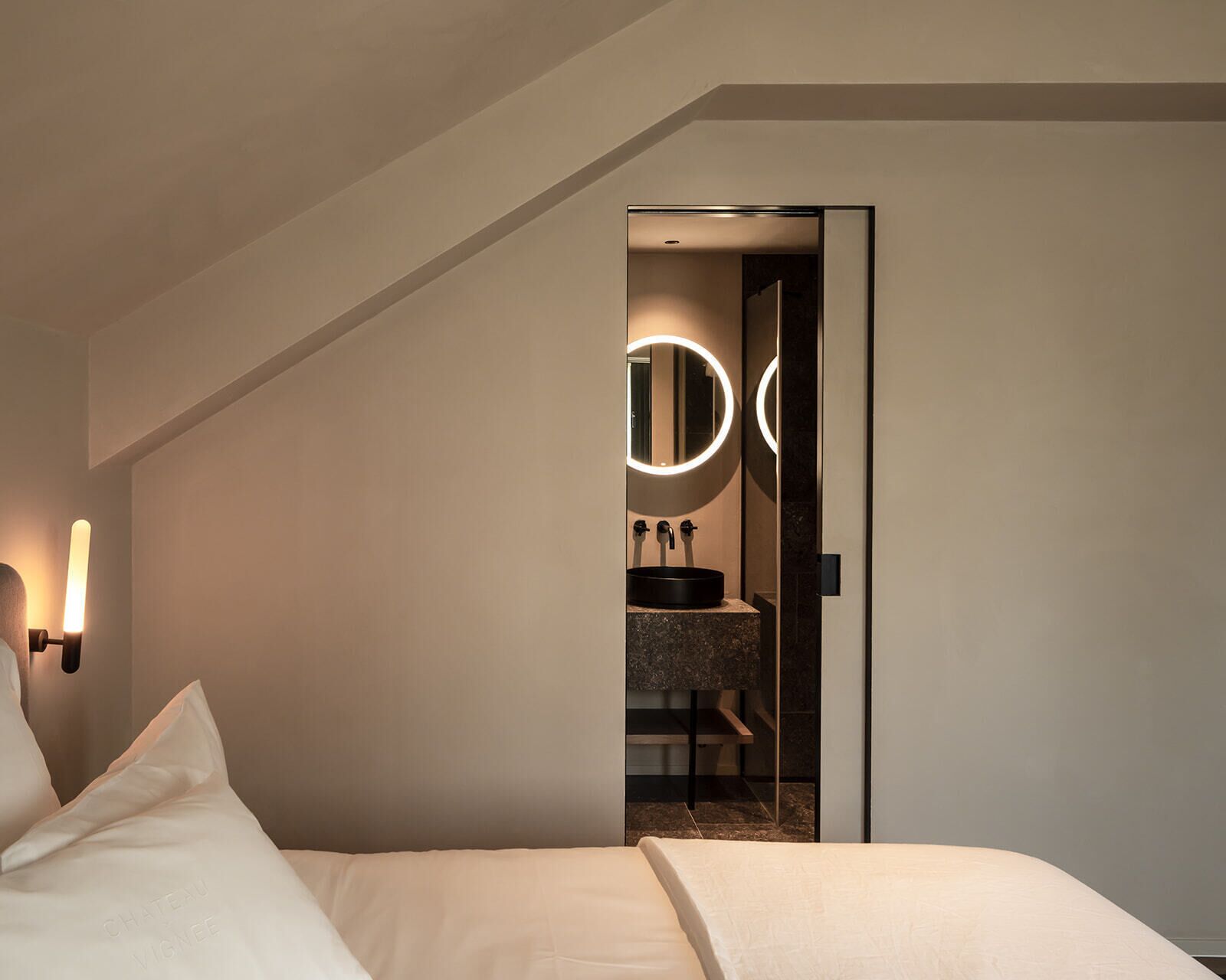 Een moderne hotelkamer met een bed, een badkamer met een ronde spiegel en een klein wastafeltje.