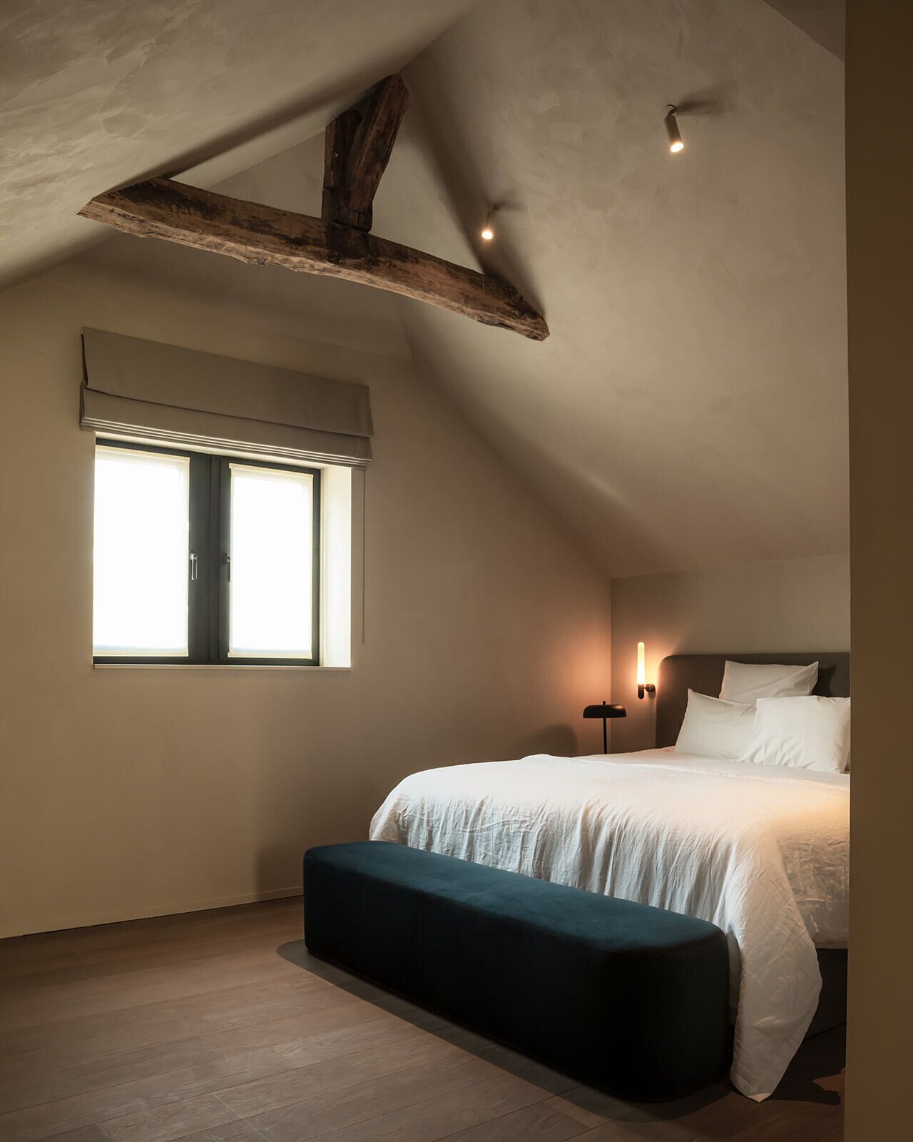 Une chambre avec un grand lit, un banc, une fenêtre et un plafond en pente avec des poutres apparentes.