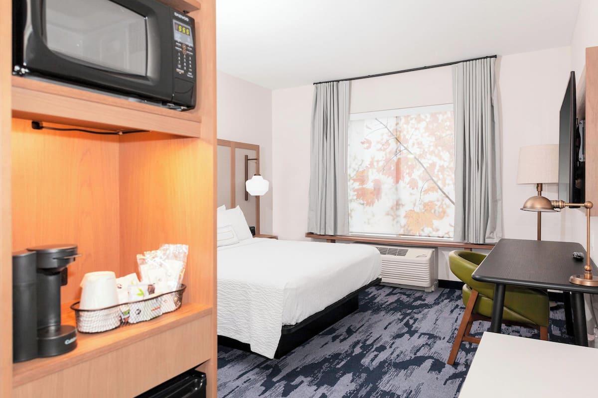 A hotel room with a bed, a desk, a microwave, a coffee maker, and a window with curtains.