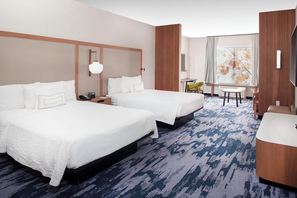 A hotel room with two beds, a desk, a chair, and a view of a tree through the window.