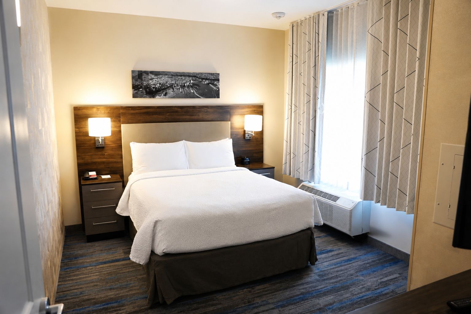 Una habitación de hotel con una cama, mesitas de noche, una lámpara de pared, una ventana con cortinas y una obra de arte colgada en la pared.