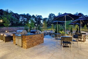 Un patio al aire libre con piso de piedra, una parrilla y mobiliario exterior con sombrillas.
