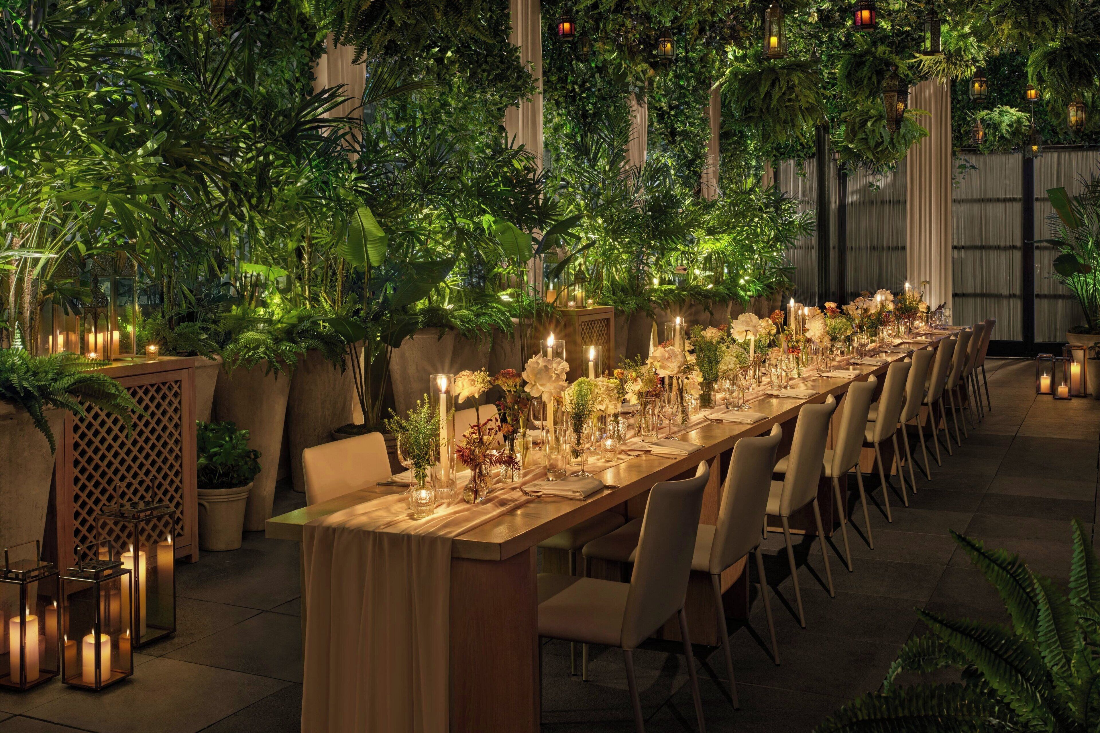 Una larga mesa de comedor dispuesta con velas y flores, rodeada de sillas, en un exuberante jardín interior con plantas y faroles.
