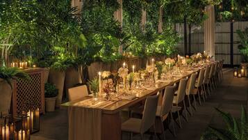 Una larga mesa de comedor dispuesta con velas y flores, rodeada de sillas, en un exuberante jardín interior con plantas y faroles.