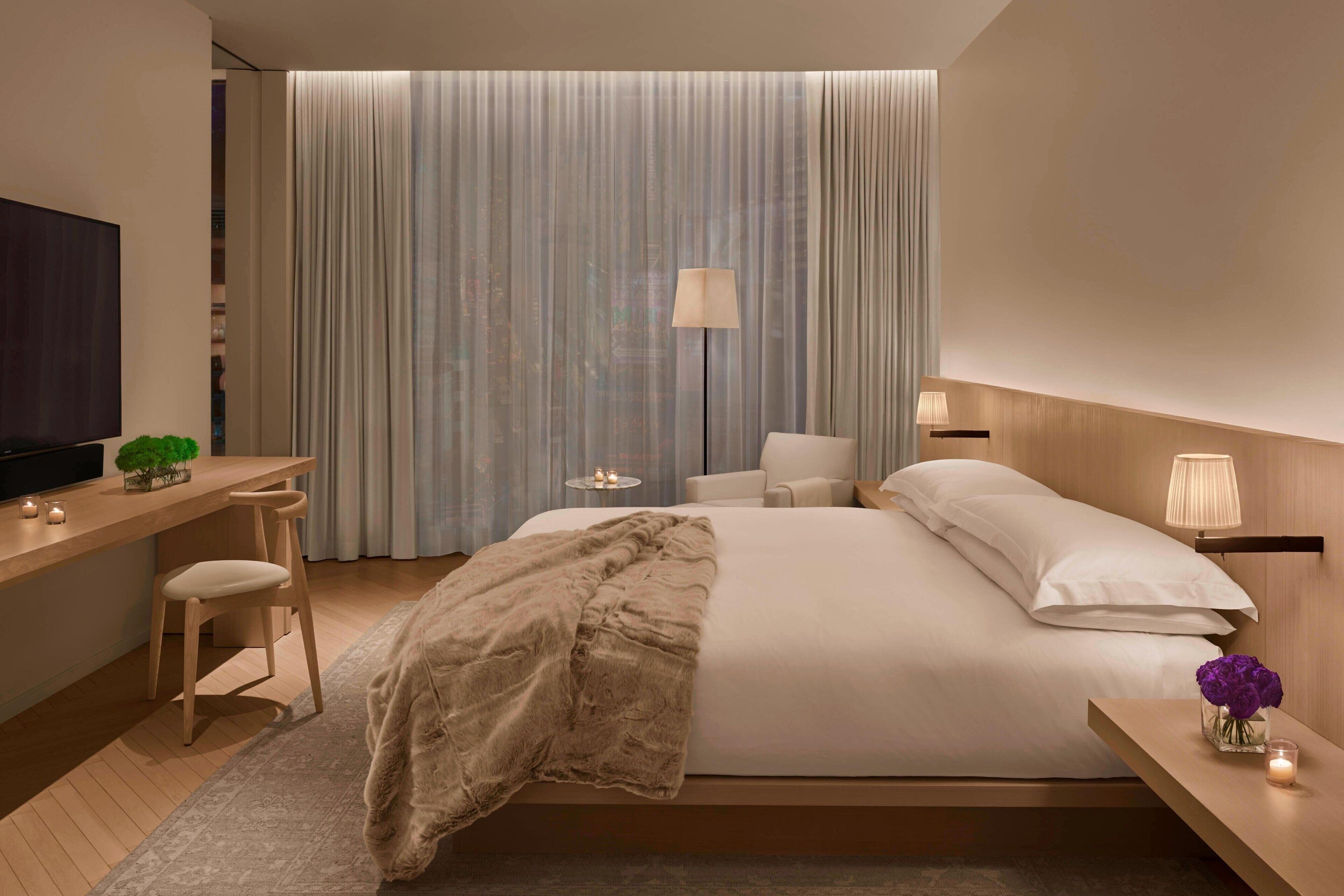 Una habitación de hotel moderna con una cama grande, un escritorio con silla, un televisor y una mesita de noche con lámpara y flores.