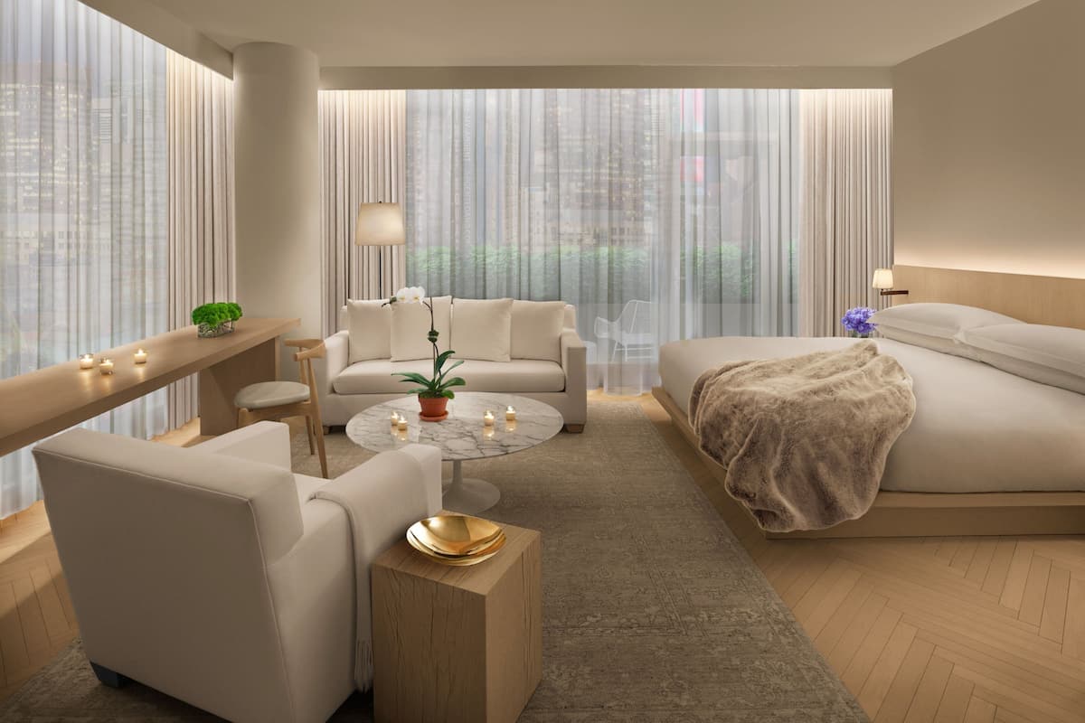 Une chambre d’hôtel moderne, dotée d’un grand lit, d’un canapé, d’une table basse et d’un bureau. Les fenêtres sont équipées de rideaux, et l’on peut admirer une vue sur la ville.