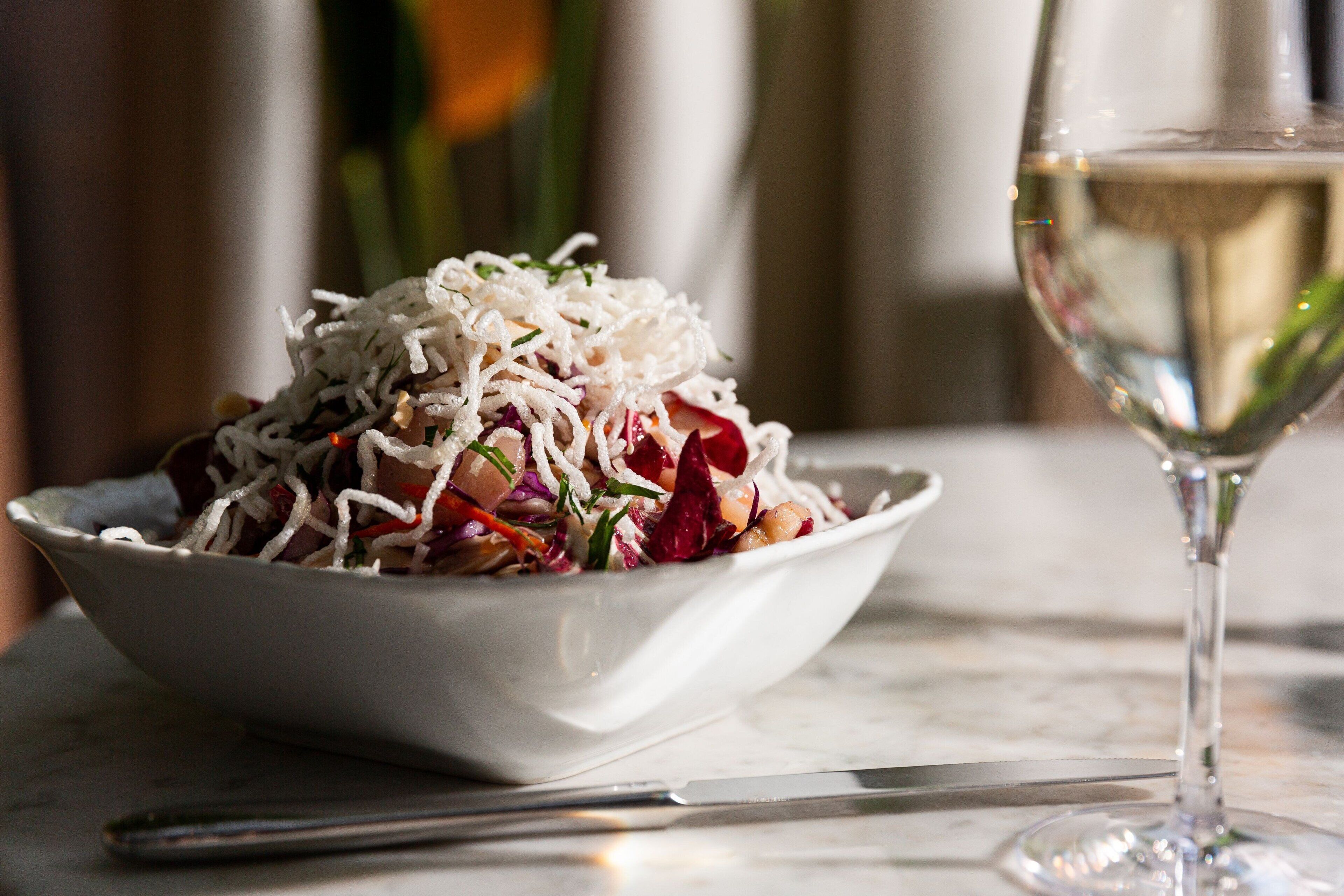 A white bowl of salad with shredded cheese, a knife, and a glass of white wine on a marble surface.
