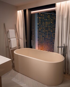 A modern bathroom with a freestanding bathtub, a wooden ladder, and a view of a building with illuminated windows.