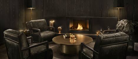 A dark-themed living room with a central fireplace, leather armchairs, a round coffee table, and two floor lamps.
