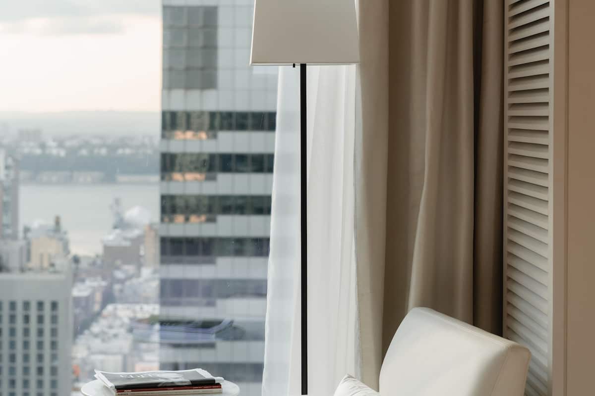 Une chambre d’hôtel moderne avec un canapé blanc, une lampe sur pied et une vue sur le paysage urbain, visible à travers de grandes fenêtres.