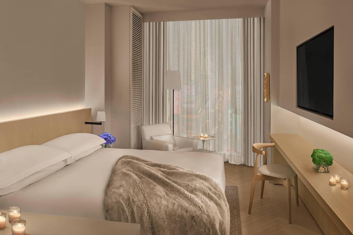 Une chambre d’hôtel moderne équipée d’un lit, d’un bureau, d’une chaise, d’une télévision et offrant une vue sur la ville grâce à de grandes fenêtres.