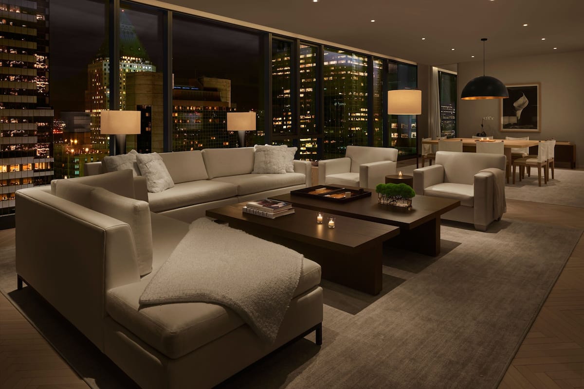 Un salon moderne avec un canapé d’angle, une table basse et un coin repas comprenant des chaises et une table. De grandes fenêtres offrent une vue sur le paysage urbain illuminé la nuit.