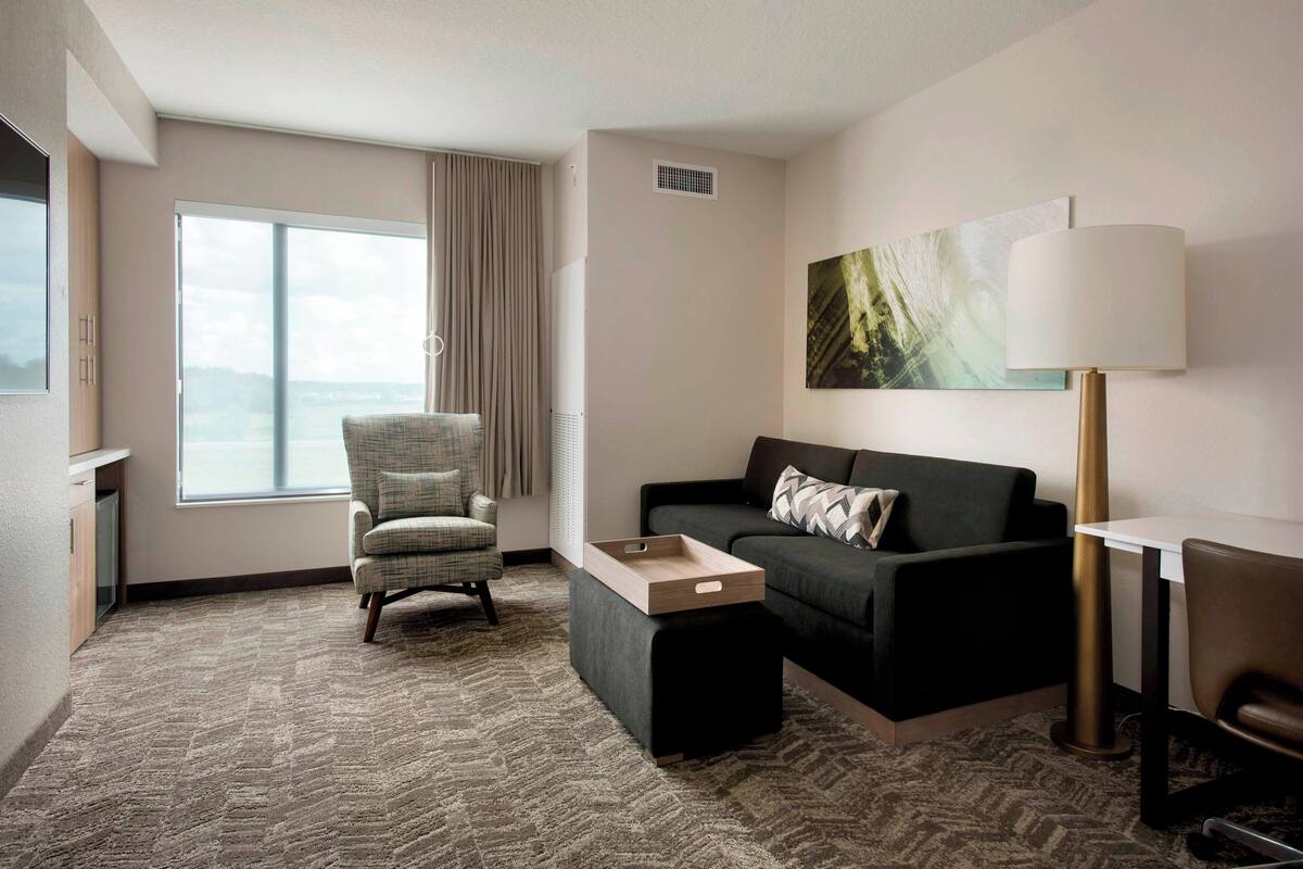 Habitación de hotel moderna con un ventanal grande, un sofá gris, una mesita de madera pequeña y una lámpara de pie.