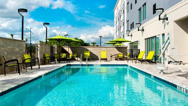 Una piscina con agua azul cristalina, rodeada de sillones de playa y sombrillas, con un edificio de amplias ventanas al fondo.