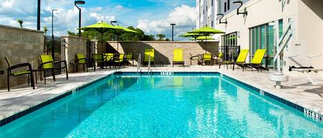 A swimming pool with clear blue water, surrounded by lounge chairs and umbrellas, and a building with large windows in the background.