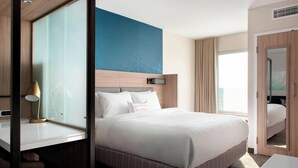 Habitación de hotel moderna con una cama grande, una mesita de noche con lámpara, un espejo amplio y vistas a la ciudad.