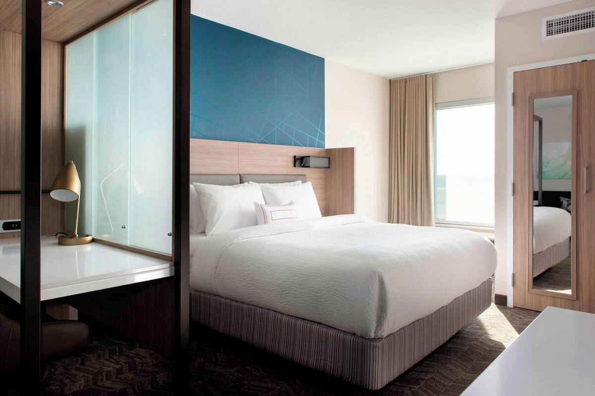 Habitación de hotel moderna con una cama grande, una mesita de noche con lámpara, un espejo amplio y vistas a la ciudad.