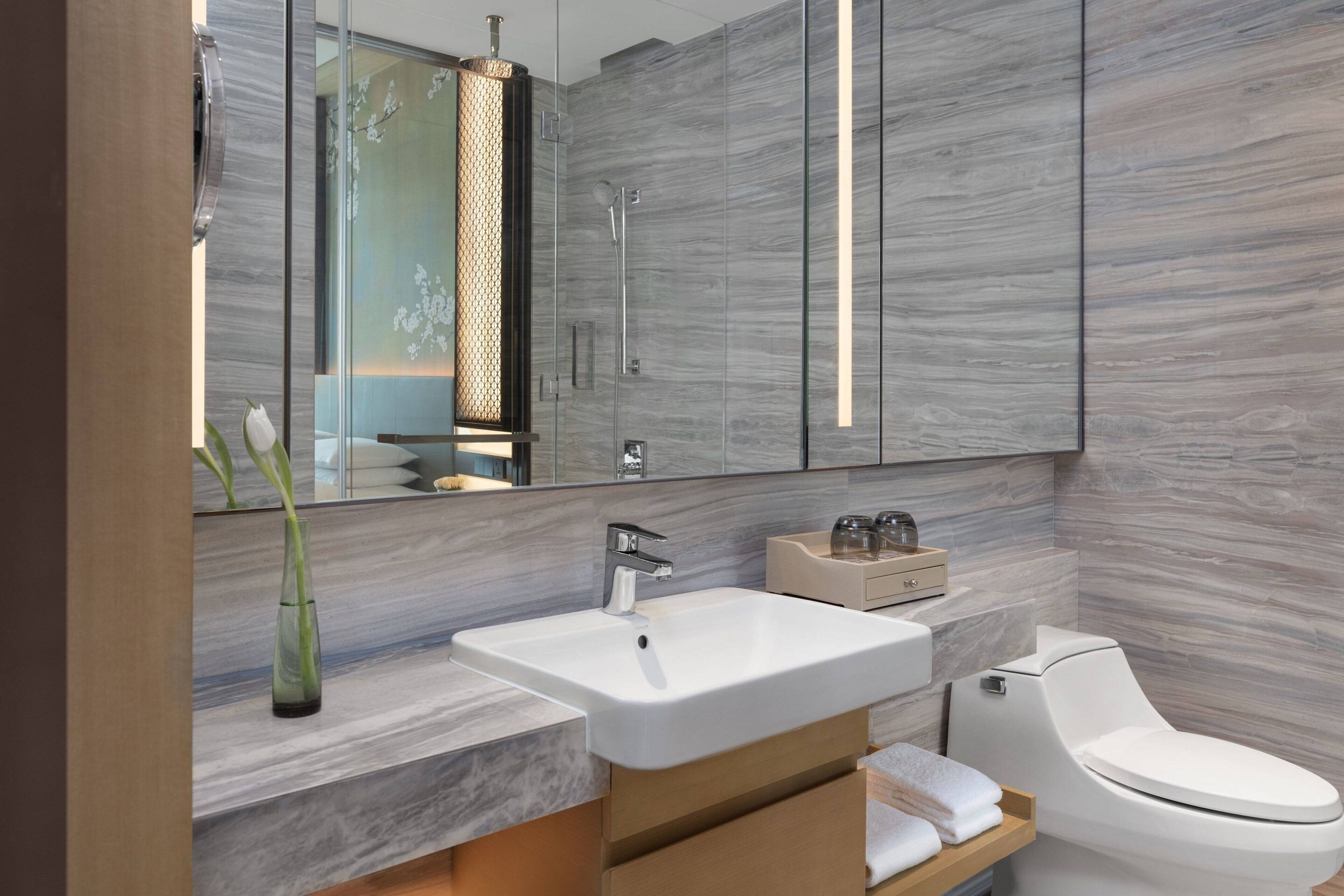 A modern bathroom with a large mirror, a white sink, a toilet, and a wooden vanity.