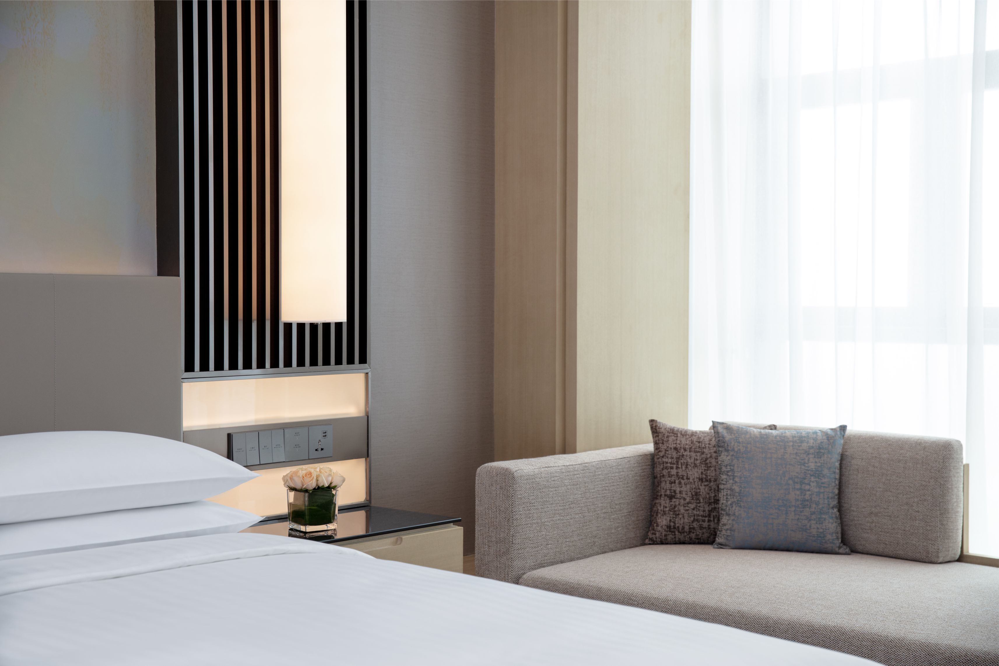 Et moderne hotelværelse med en seng, en sofa, et sengebord med en vase med blomster, og et stort vindue med lette gardiner.
