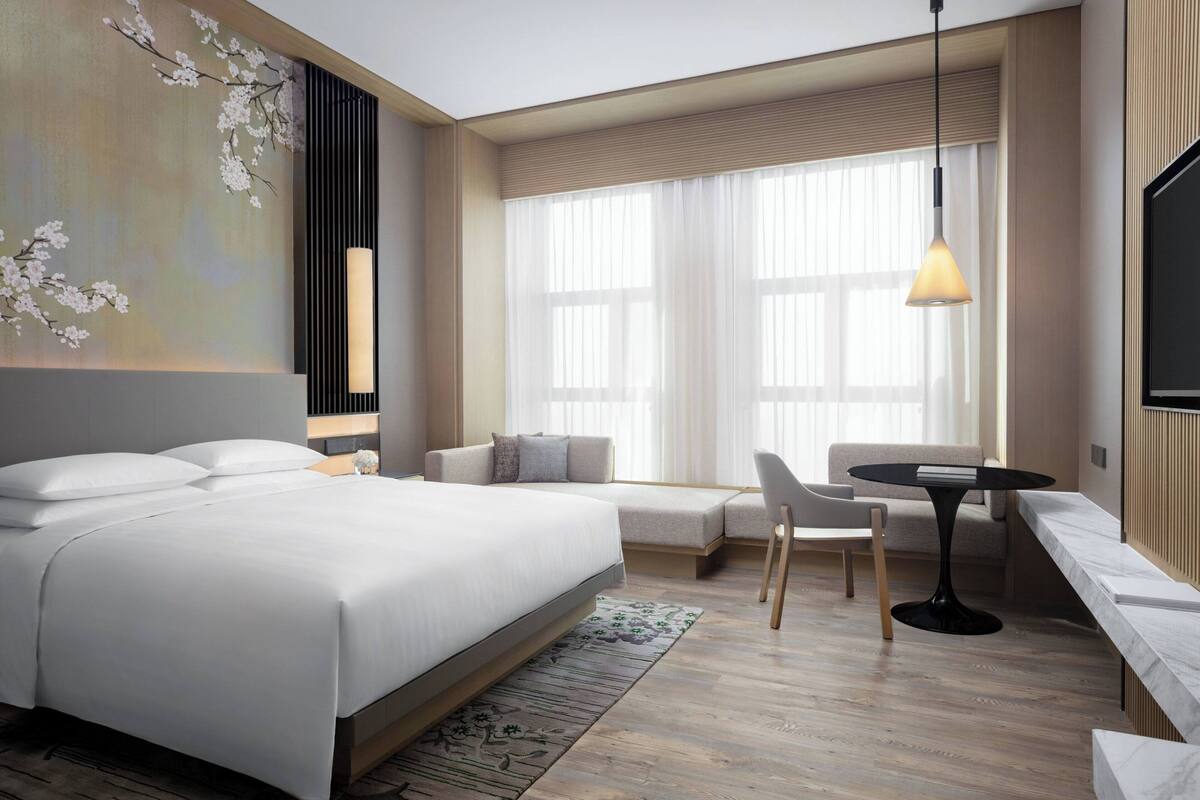 A modern hotel room with a large bed, a seating area with a sofa and chairs, a small table, and a large window with curtains.
