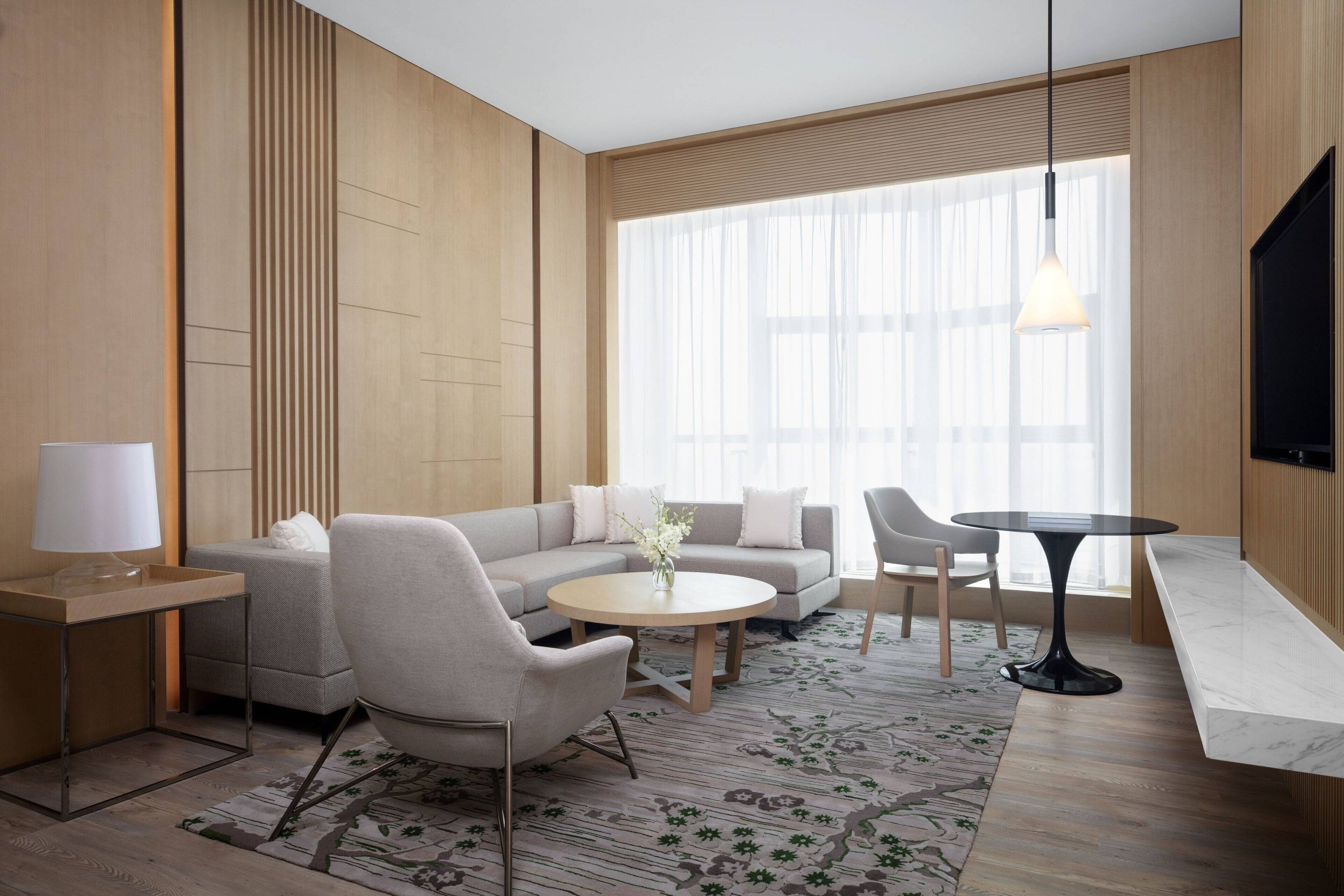 A modern hotel room with a sectional sofa, a round coffee table, a side table, and a TV mounted on the wall.