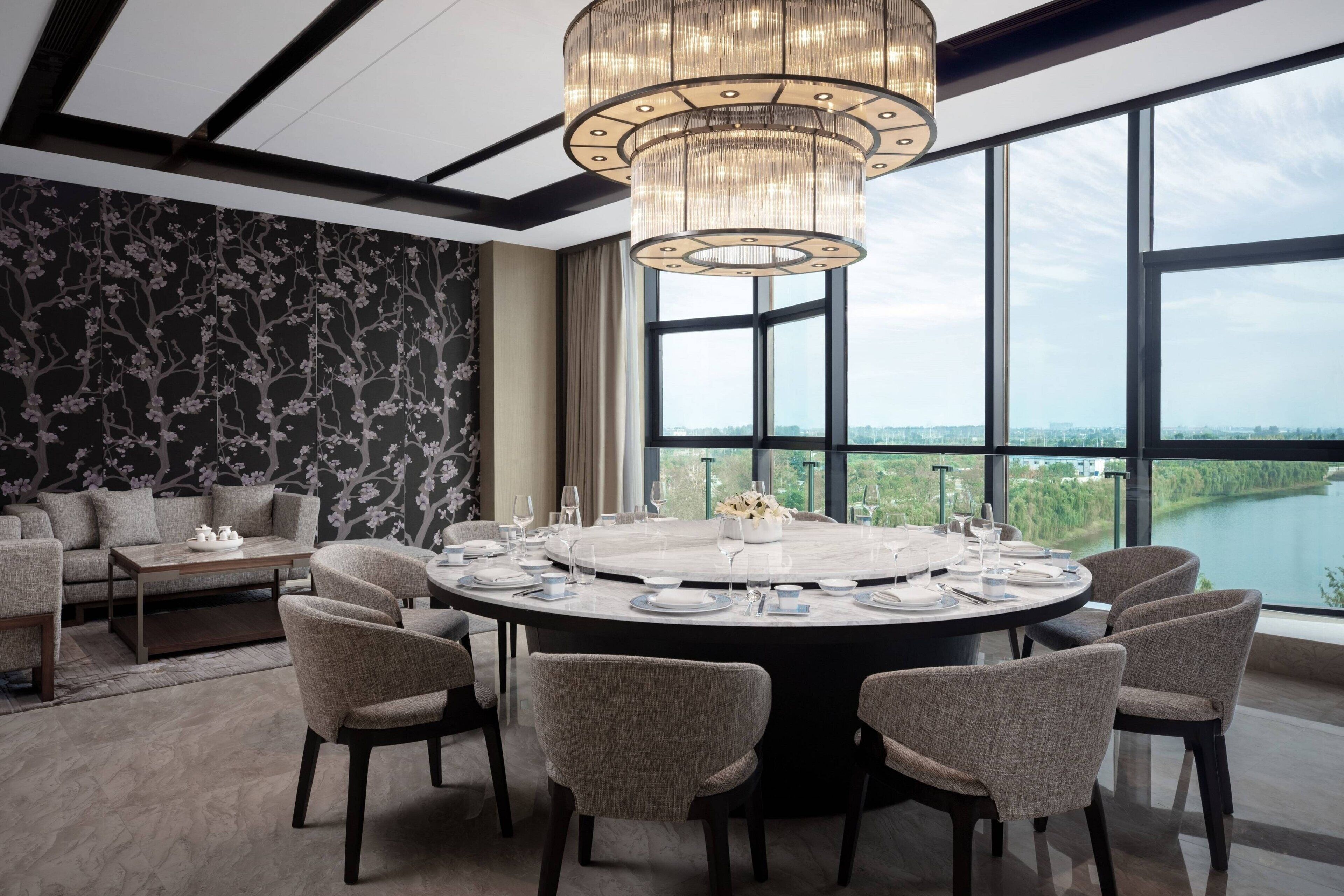 A modern dining area with a round table set for six, surrounded by six chairs. A large chandelier hangs from the ceiling, and the room features large windows offering a view of a body of water.